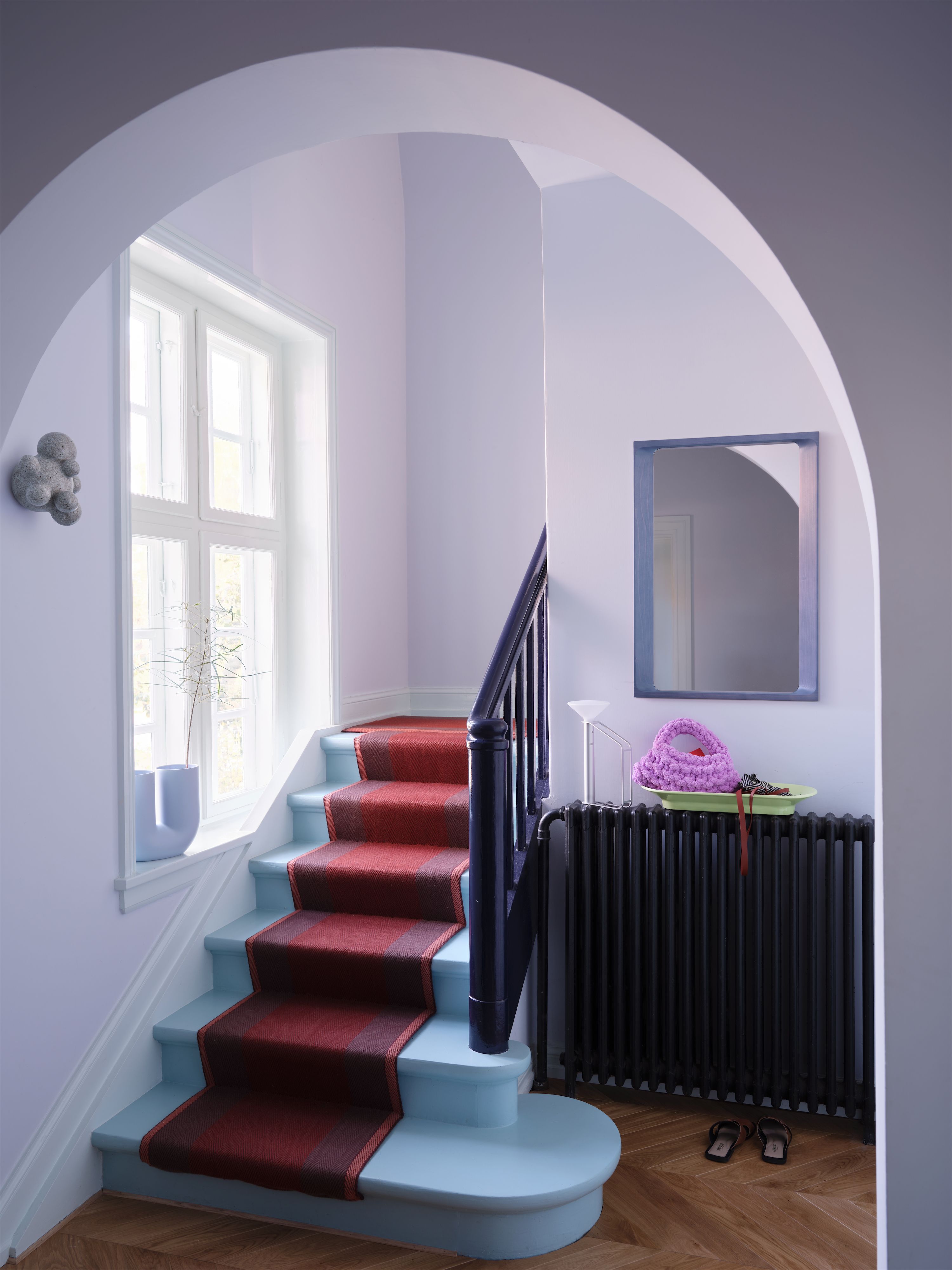 Treppenhaus mit fliederfarbenen Wänden, blau gestrichener Treppe mit rotem Läufer und Wandspiegel.