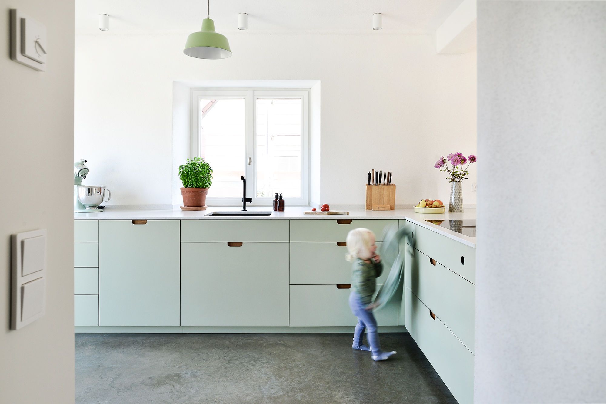 Moderne Linoleum-Küche in Mintgrün mit Griffmulden von Nuance Living, in der ein Kind spielt - robustes Design für den Familienalltag.