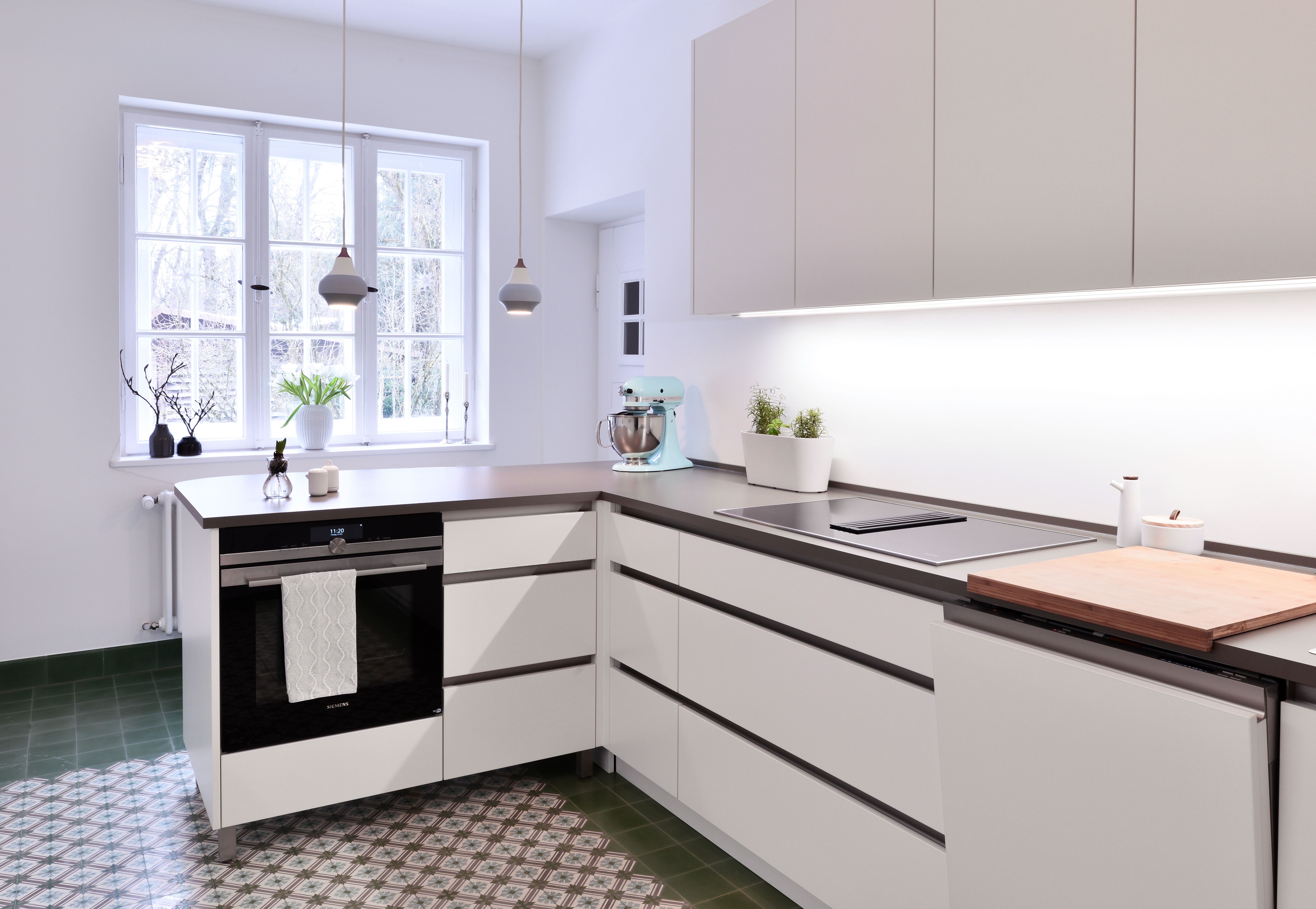 Moderne, weiße L-Küche von Nuance Living mit grifflosen Fronten und dunkler Arbeitsplatte, kombiniert mit Vintage-Bodenfliesen in einem hellen Altbau-Raum.