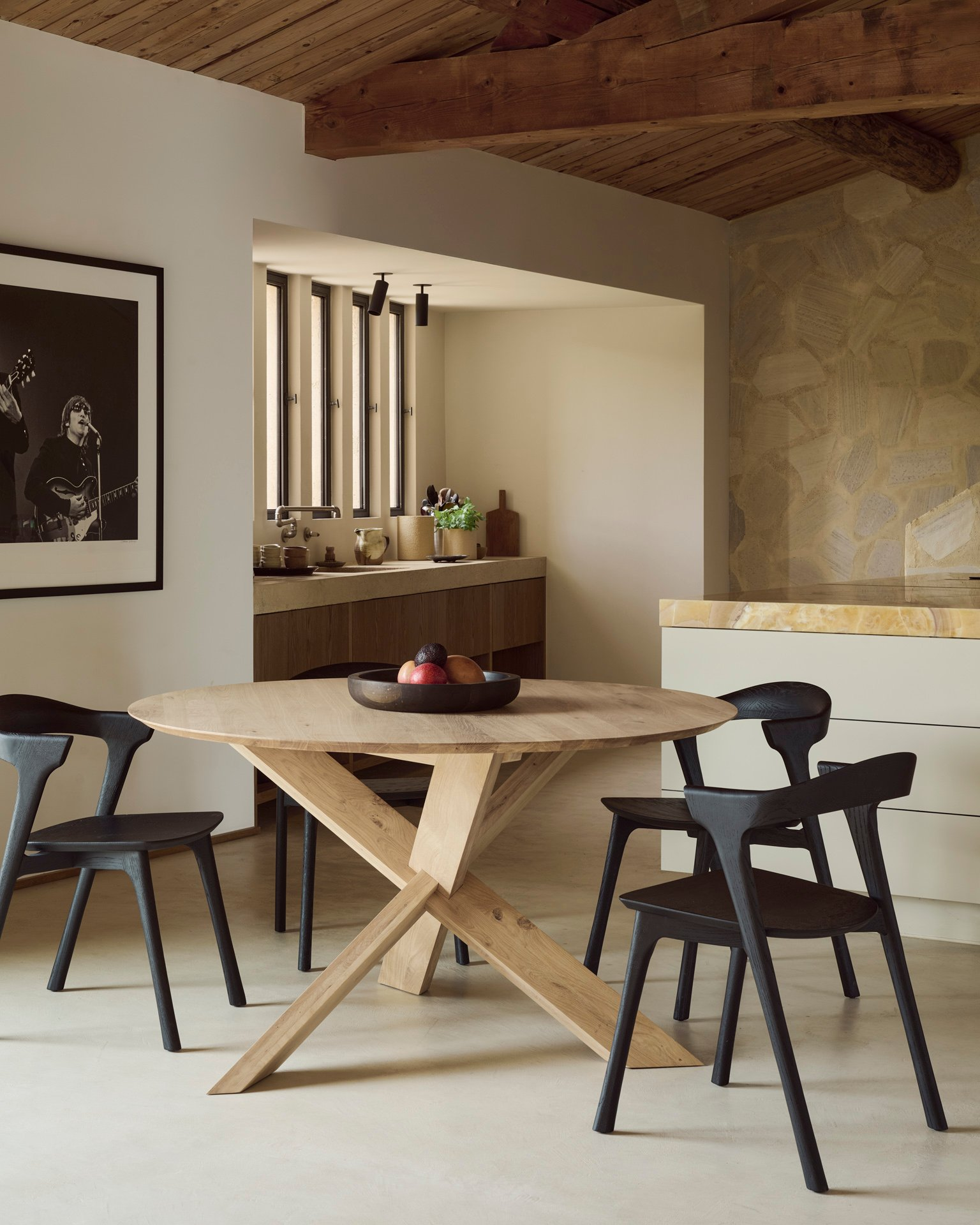 Heller Essbereich mit rundem Massivholztisch, schwarzen Design-Stühlen und einer rustikalen Steinwand im Hintergrund